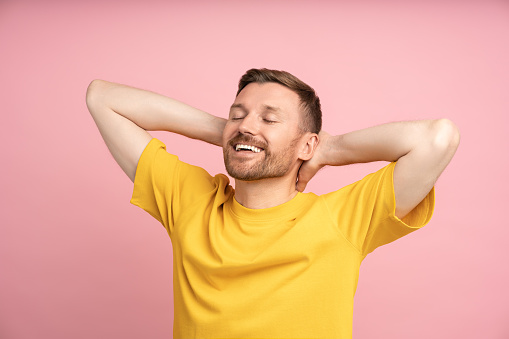 Relaxed man holding hands behind head smiling with closed eyes standing on pink background. Satisfied resting handsome guy with beard stubble enjoying pastime. Dreaming calm mood, optimism concept.