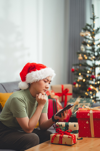 Asian woman sitting sofa in living room decorated with a Christmas tree looking at smartphone screen and having problem feel worried about spam or bad news.