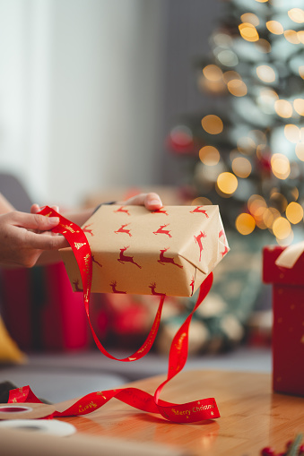 Close-up shot of woman tying ribbon on gift box presents for Christmas, sitting in living room decorated with a Christmas tree.