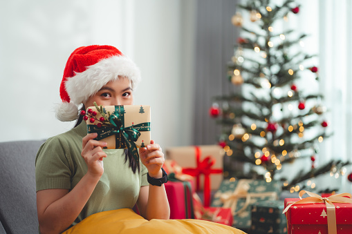 Asian woman sitting in living room decorated with a Christmas tree, smiles happily and showing Christmas present to camera.