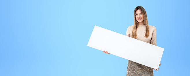 Young smiling woman holding a mockup blank signboard in hands, empty copy space blue background. Concept of recommendation, announcement and advertisement