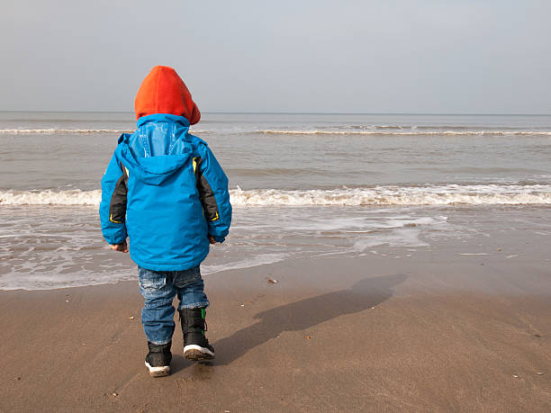 small boy in winter clothing stands alone on a sandy beach looking out to sea