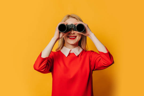 Stylish blonde hair woman in red dress with binoculars on yellow background stock photo