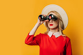 blonde hair woman in hat and red dress with binoculars on yellow background