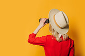 Back side view on woman in hat and red dress with binoculars on yellow background