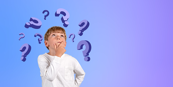 Portrait of thoughtful young schoolboy in casual clothes standing with hand on chin near blue wall and looking at question marks. Concept of curiosity, education and choice. Copy space