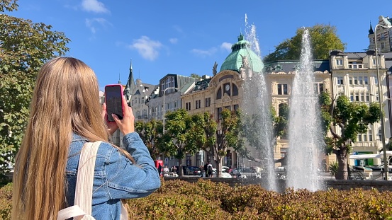 Stylish woman traveler with a backpack walks and takes photos of a fountain in the Old Town in Europe.
