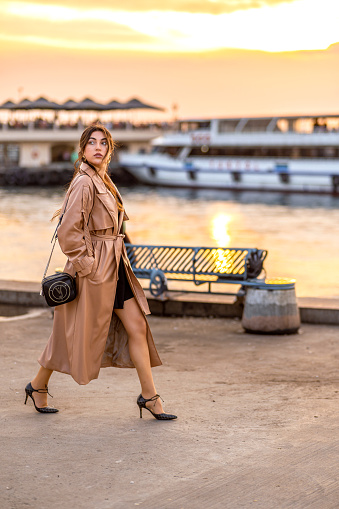 Young beautiful woman in trench coat walks along the beach in the city