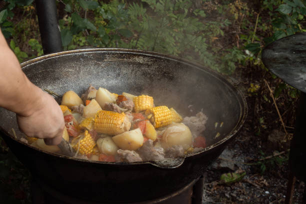 Large pieces of meat, ears of corn, potatoes and vegetables are fried in a cast-iron cauldron on an outdoor wood-burning stove. Street food