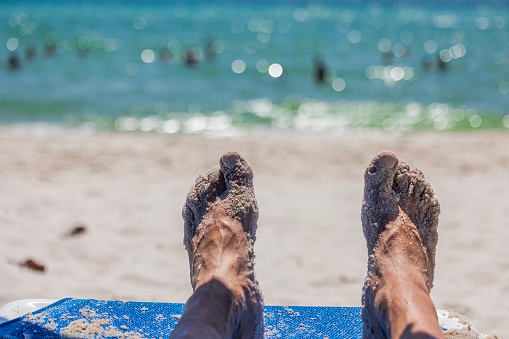 Close-up of men's feet on sunbed with focus on foreground and blurred background of Atlantic ocean water. Miami Beach, USA.