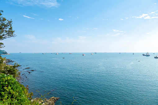 Fishing boats on the horizon at the sea. The water is blue, the sky is a bit cloudy and blue. The boats are far in the distance. There are some rocks at the sea shore with green vegetation. The picture was made in October in the bay of Busan city, South Korea.