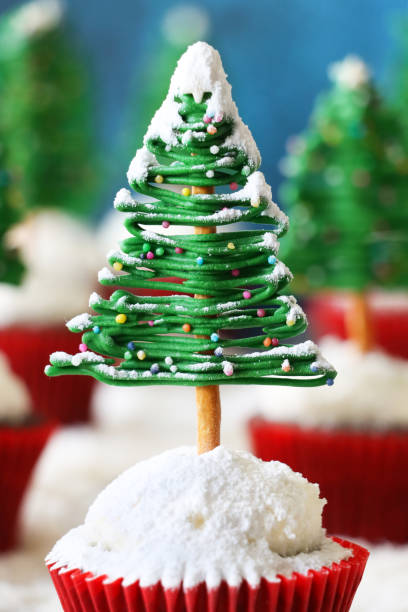 istock Full frame image of batch homemade, Christmas tree design chocolate cupcakes, buttercream coated in desiccated coconut topping, white chocolate and pretzel stick trees, displayed in snowy, conifer forest scene, blue background, focus on foreground 1789404363