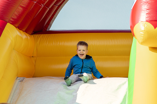 Delighted little boy sitting on inflatable trampoline and smiling. The child wants to slide down the big inflatable slide in the amusement park.