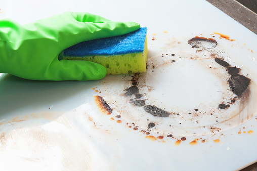 A man is cleaning a modern white electric stove wearing green rubber gloves. Cleaning an induction cooker with a yellow and blue sponge.