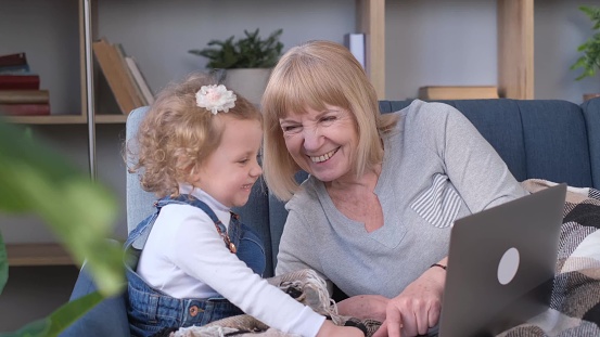 Smiling grandmother and little granddaughter using laptop together, sitting on cozy sofa in living room, watching video or cartoons, having fun on weekend.