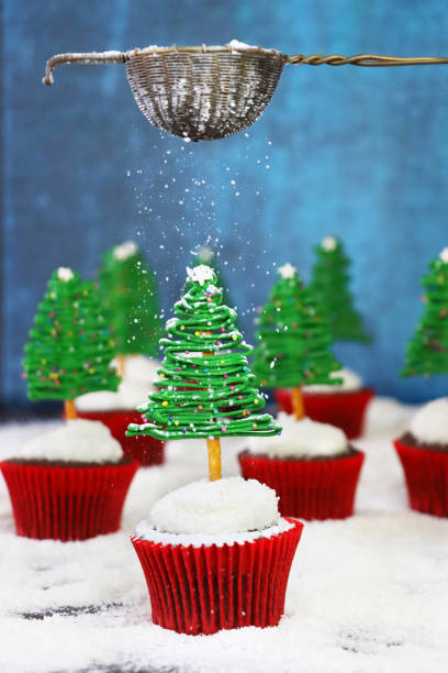 istock Image of icing sugar sifted over batch homemade, Christmas tree design chocolate cupcakes, buttercream coated in desiccated coconut topping, white chocolate and pretzel stick trees, displayed in snowy, conifer forest scene, blue background 1789442063