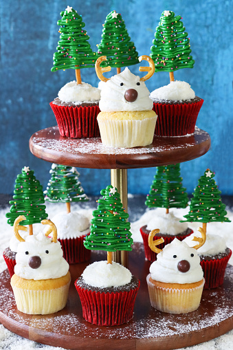 Stock photo showing close-up view of a cake stand displaying a batch of Christmas tree and reindeer design cupcakes, in paper cake cases, displayed against a blue background.