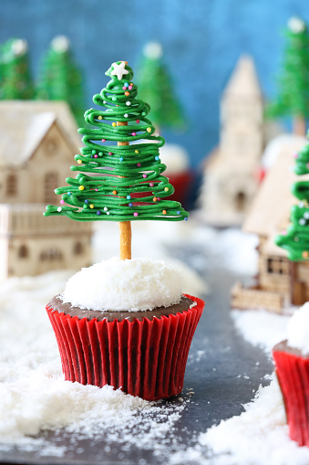 Stock photo showing close-up view of a batch of freshly baked, homemade Christmas tree design chocolate cupcakes, in red metallic paper cake cases, displayed in a snowy, Christmas village scene. The cupcakes are topped with butter icing coated in desiccated coconut from which protrude Christmas trees made from iced white chocolate, coloured with green food dye, on trunks of salted, pretzel sticks. The trees have been decorated with multicoloured sugar sprinkles. Home baking concept.