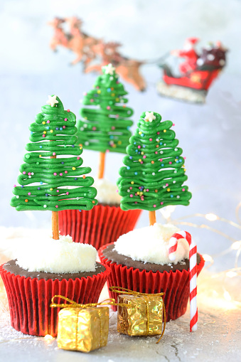 Stock photo showing close-up view of three Christmas tree design chocolate cupcakes, in red metallic paper cake cases, displayed against a grey mottled background. The cupcakes are topped with butter icing coated in desiccated coconut from which protrude Christmas trees made from iced white chocolate, coloured with green food dye, on trunks of salted, pretzel sticks. The trees have been decorated with multicoloured sugar sprinkles. Home baking concept.