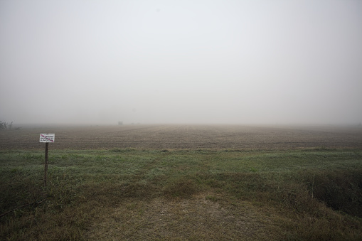 Entrance  to a field with a sign over a trench on a foggy day in the italian countryside