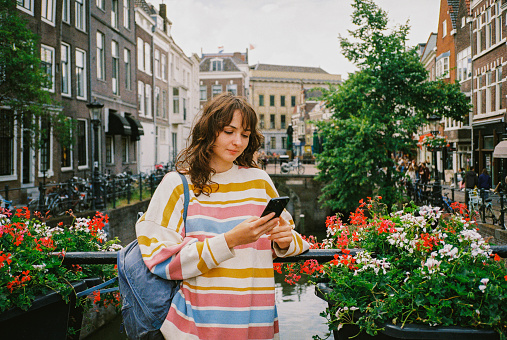 Cheerful woman with backpack standing in Utrecht in spring and using smartphone
