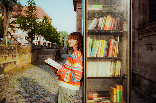 Woman reading book from book crossing shelf in the city. Shot on camera film