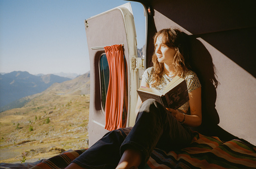 Cheerful woman reading book in camper van in mountains