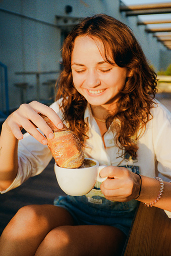 Cheerful woman dipping croissant in cappuccino sitting on sun