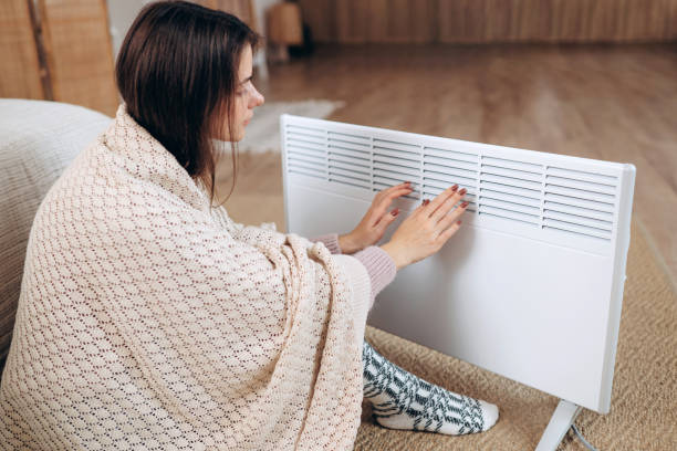 istock Cold home, freezing. Using heater at home in winter. Woman warming her hands sitting by device and wearing warm clothes. Heating season. 1789764849