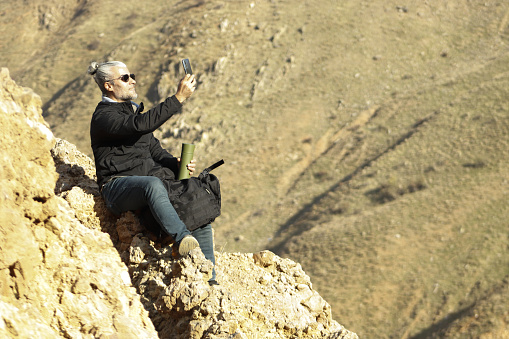 Vigorous man with sunglasses and backpack, climbing a mountain on a sunny day, walking outdoors, climbing, sitting, resting, drinking water from a thermos