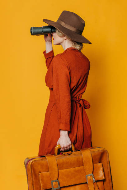 Back side view on woman in hat and brown dress with suitcase and binoculars on yellow background