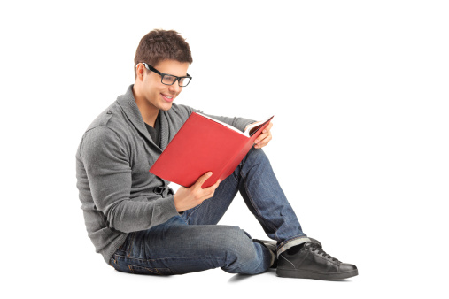 Smiling guy sitting on a floor and reading a book isolated on white background