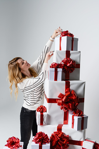 A gorgeous beautiful young woman poses among a mountain of white gift boxes in a gray background. Beautifully decorated for Christmas. Holiday fashion. Sales and shopping.