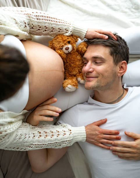 The parents lying on the bed, with the proud father listening to the naked, round baby bump and looking excitedly at it. Last month of pregnancy - week 39. White background. Description: The parents lying on the bed, with the proud father listening to the naked, round baby bump and looking excitedly at it. Last month of pregnancy - week 39. White background. bear stomach stock pictures, royalty-free photos & images