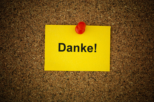 A yellow paper note with the word Danke! (Thank you in German) on it pinned to a cork board. Close up.