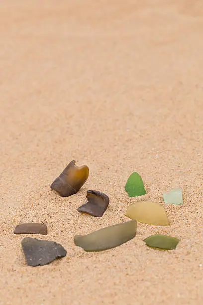 A grouping of 8 pieces of different colored seaglass is photographed in the sand.  Atlantic seaglass featured is brown, dark green, light green, and pale green.