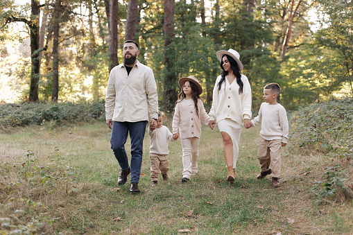 Young mother, father with daughter and sons are walking, having fun in autumn forest. Family holding hands enjoying time together at background of trees. Happy parenthood and childhood concept