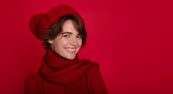 Close up portrait of smiling woman in casual red sweater and winter hat and scarf in good mood isolated on bright red background. Advertising female studio portrait. Christmas time