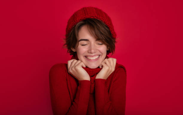 Close up portrait of smiling woman in casual red sweater and winter hat and scarf in good mood isolated on bright red background. Advertising female studio portrait. Christmas time stock photo