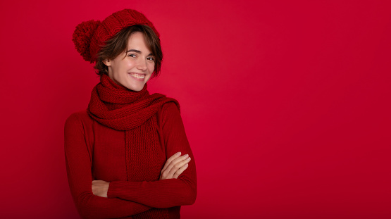 Close up portrait of smiling woman in casual red sweater and winter hat and scarf in good mood isolated on bright red background. Advertising female studio portrait. Christmas time