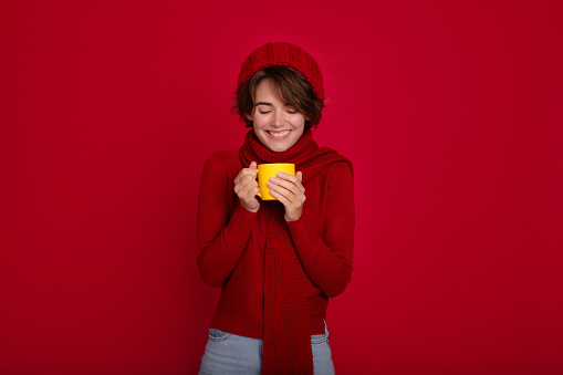 Close up portrait of young smiling woman wearing a winter hat, sweater and scarf with a yellow mug with tea, coffee or cocoa in hands isolated on red background. Warm drinks.
