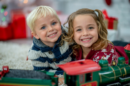 A sweet little girl and her younger brother play with a train on Christmas morning.  They are both wearing warms sweaters and are smiling.