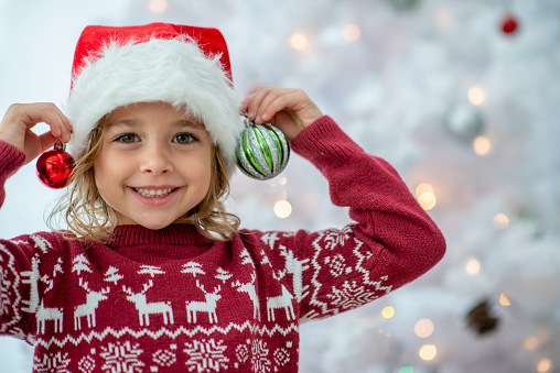 A sweet little girl in a red Christmas sweater and a Santa hat, holds up two decorations as she prepares to adorns the tree.