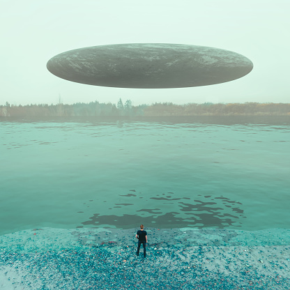 The image portrays a surreal and mysterious scene. It features an individual standing at the edge of a body of water, possibly a lake, with their back to the viewer. The person appears to be looking at a large, oval-shaped object hovering above the water, which has the characteristic appearance of a UFO.