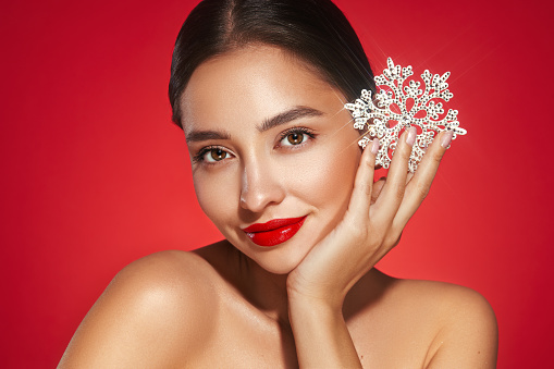 Beautiful young woman with red lipstick, shiny clean skin and face, showing glowing snowflake, near face and smiling, isolated on a red background. Christmas holidays concept