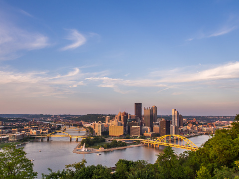 As the sun sets over Pittsburgh, the city's captivating skyline, Point State Park, and the iconic yellow bridges spanning the Allegheny and Monongahela Rivers create a picturesque scene against the backdrop of a stunning evening sky. This serene sunset view beautifully encapsulates the city's charm and natural beauty.