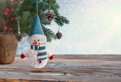 Christmas composition with a happy snowman on an empty wooden table. Christmas decoration in the form of a snowman on a Christmas tree branch. Defocused Christmas lights in the background. Christmas and New Year holiday background or banner.