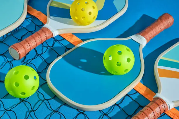 Photo of pickleball paddles with balls on top of a net on a blue background illuminated with hard light