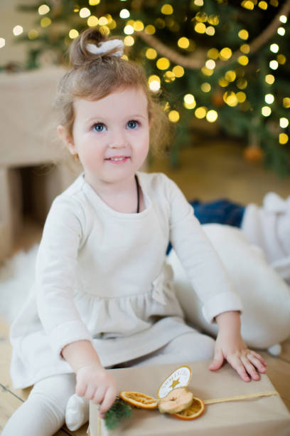 Cute little girl in a white dress sits near Christmas tree with decorations and lights and plays with gift boxes