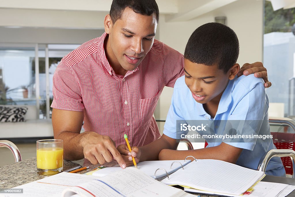 Father Helping Son With Homework In Kitchen Father Helping Son With Homework In Kitchen Reading Question With Arm Round Son Father Stock Photo